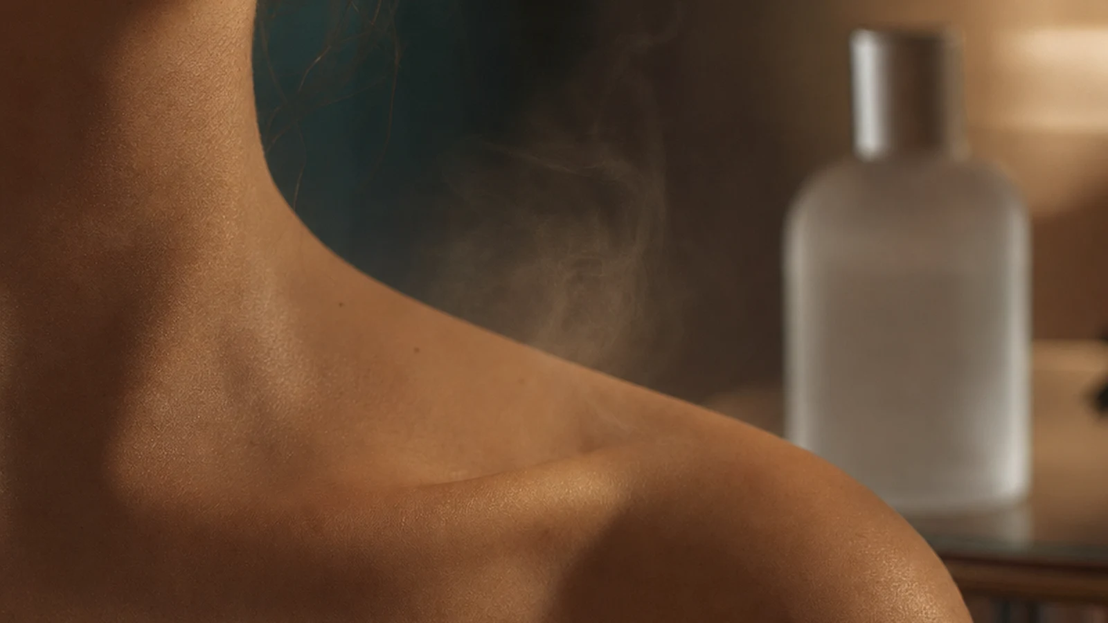 A woman's collarbone and shoulder in warm low light, a faint haze of scent lifting from the skin, with an unlabelled frosted-glass fragrance bottle out of focus behind